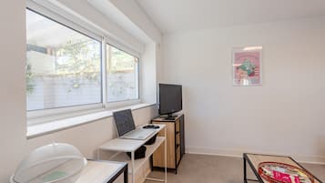 Una habitación con escritorio, laptop, televisión, ventana con vistas a la vegetación y un cartel en la pared.