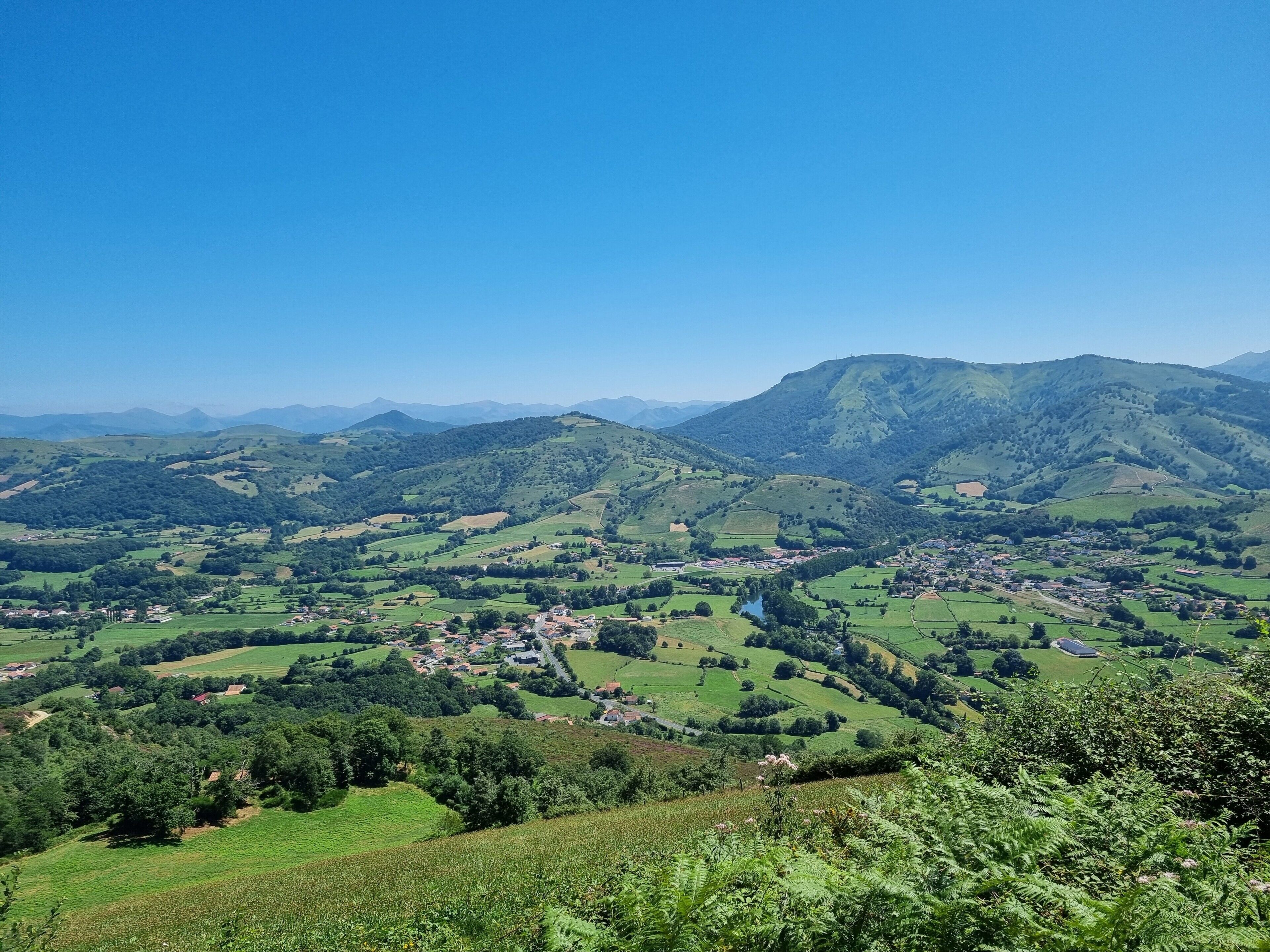 Una vista panoramica di una valle con dolci colline, un piccolo paese e montagne in lontananza.
