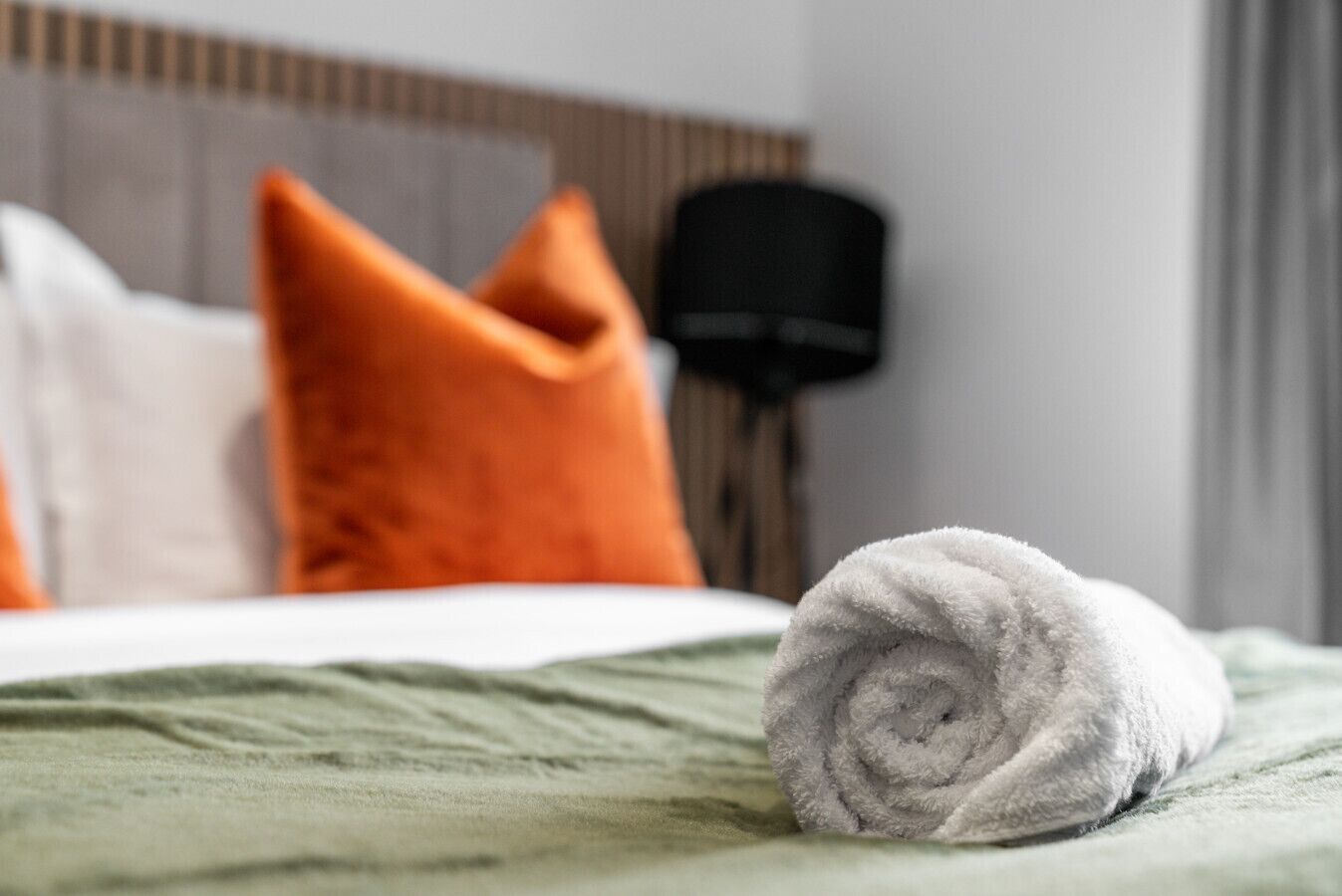 Cama bem arrumada com uma toalha enrolada, um edredom verde e duas almofadas, uma laranja e outra branca, em um quarto de hotel.