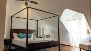 Una cama con dosel, un ventilador de techo, una mesita de noche, una silla y un amplio ventanal con cortinas.