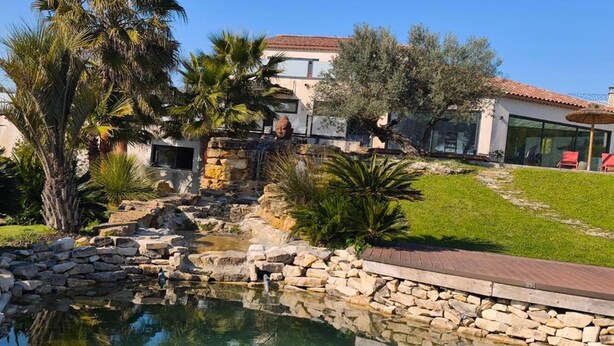 Una piscina con paredes de piedra, palmeras y una casa con techo rojo al fondo.