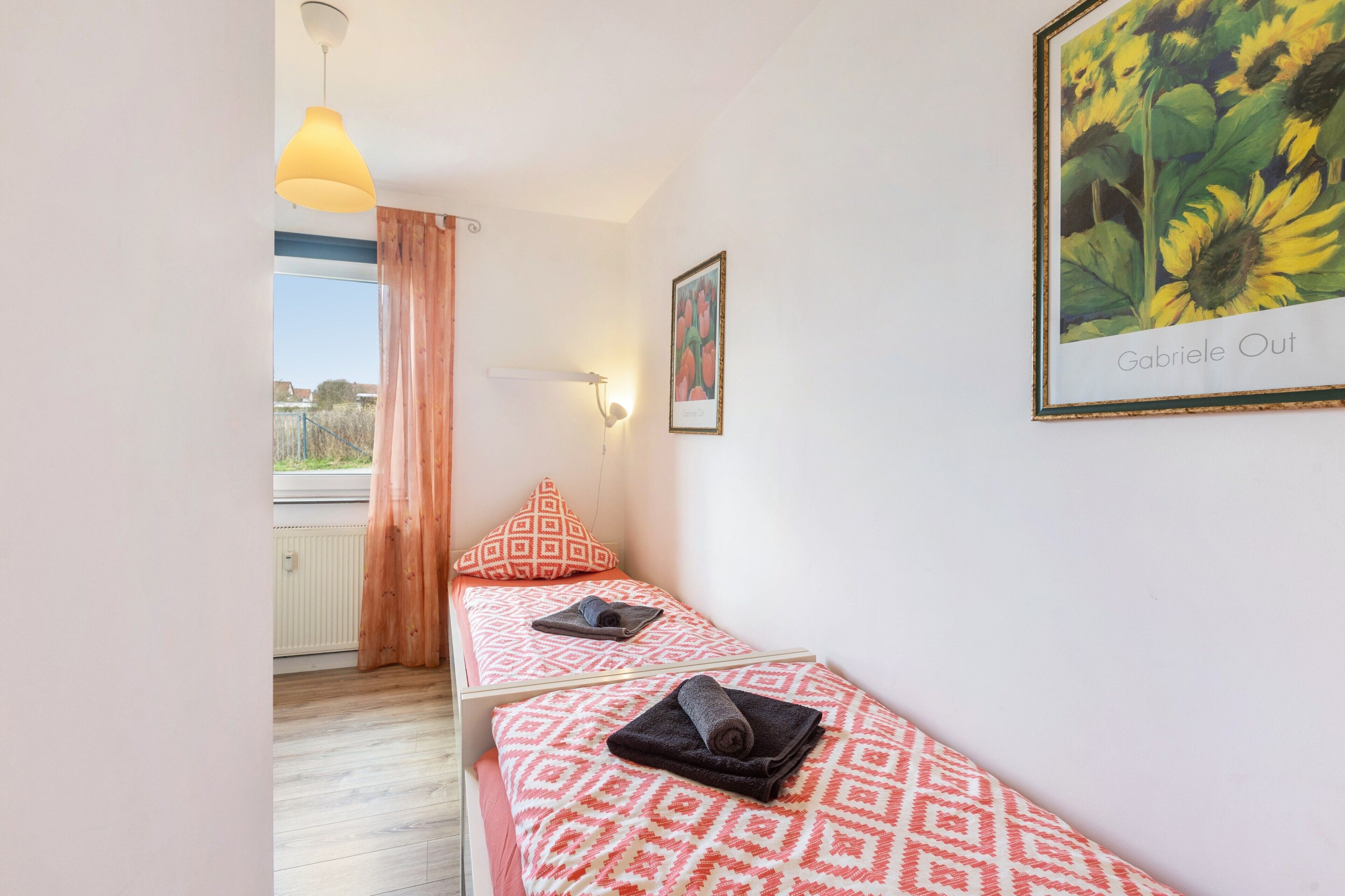 Een kamer met twee bedden, een raam met gordijnen, een wandlamp en een lijst met zonnebloemen.