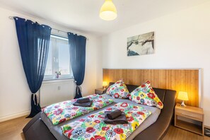 Ett sovrum med två sängar, en sänggavel i trä, ett nattduksbord med en lampa, ett fönster med gardiner och en tavla på väggen.