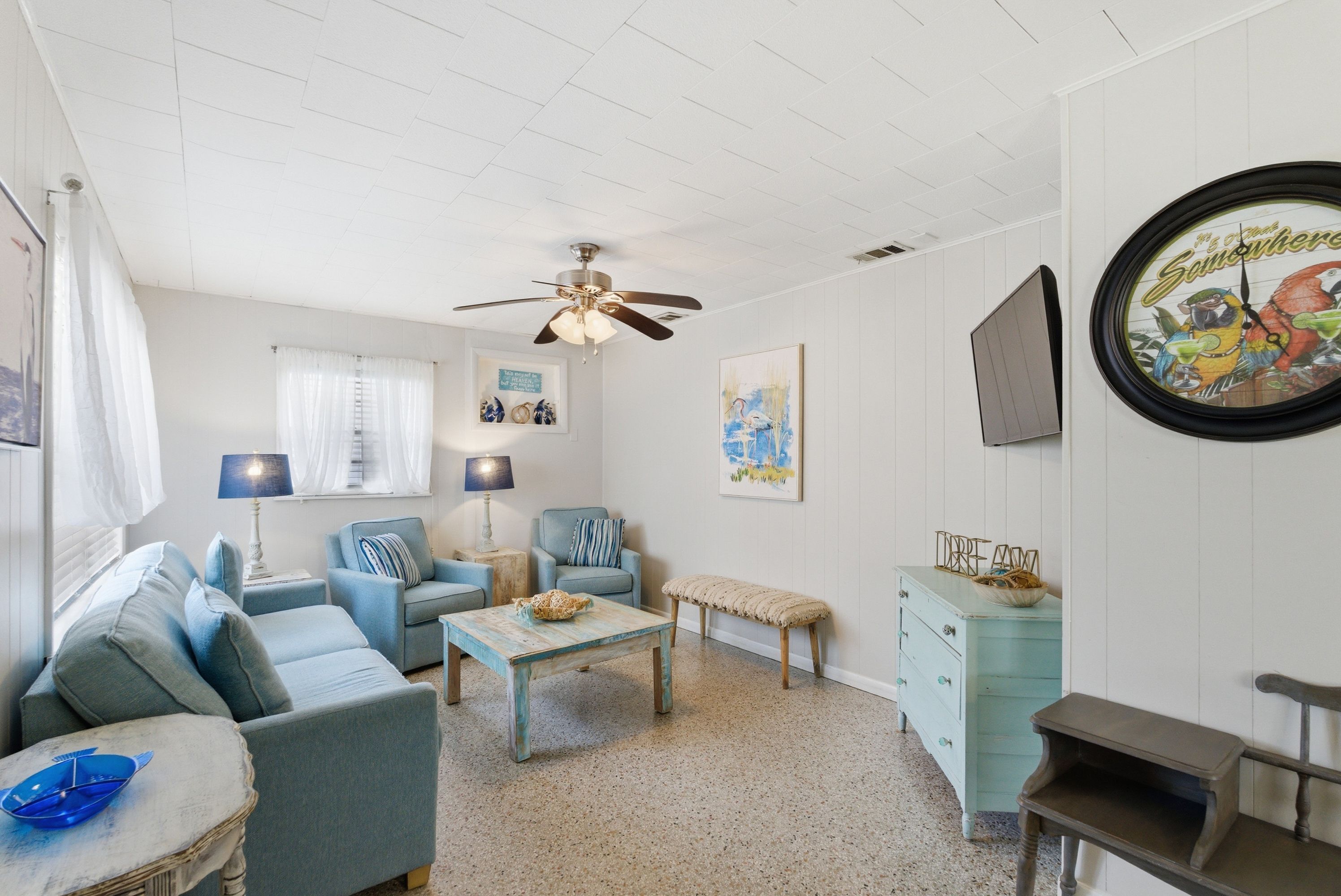 Une pièce avec un ventilateur de plafond, une horloge, un canapé bleu, une table basse et une commode.