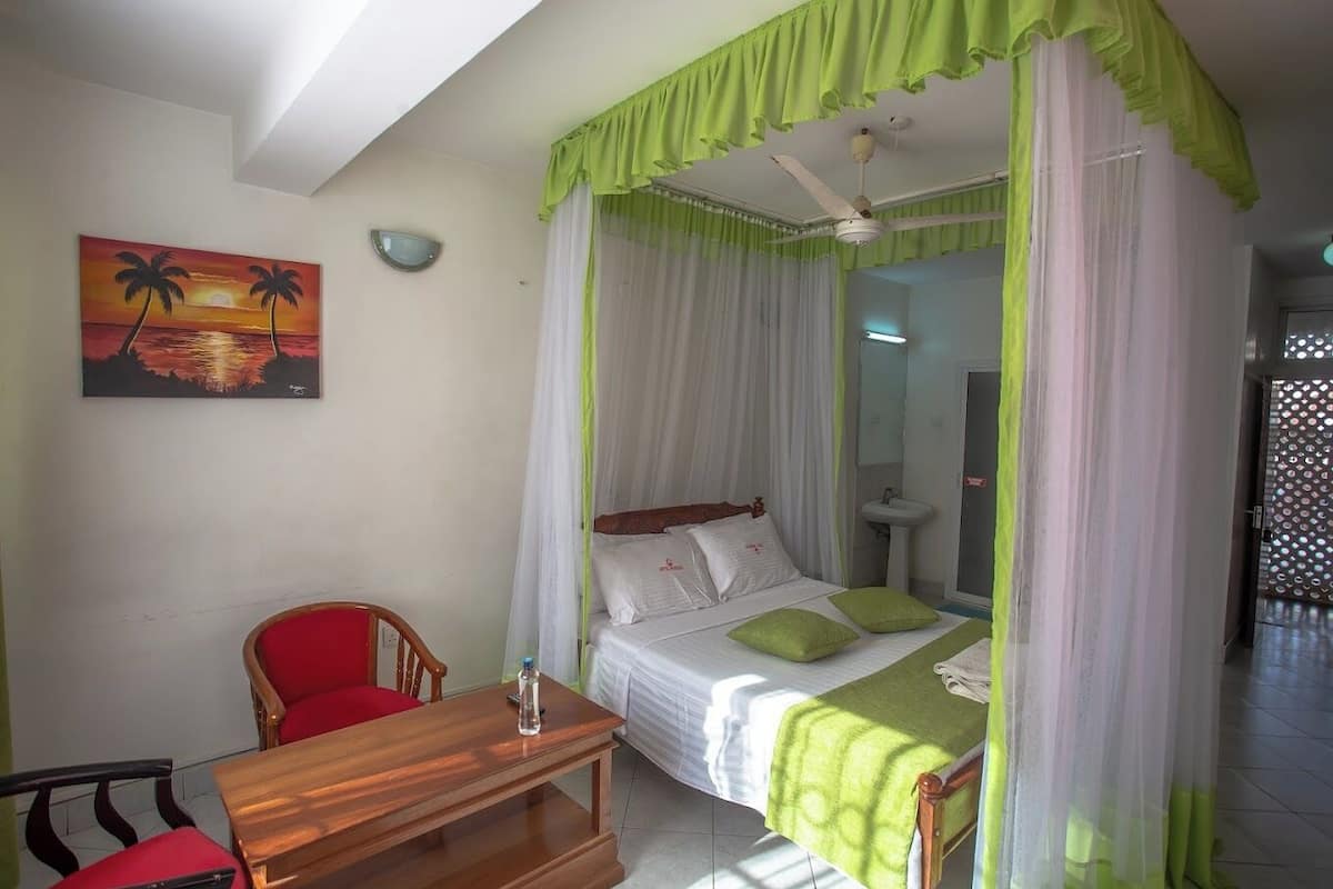 A room with a bed under a green canopy, a wooden table with a chair, a painting of a sunset, and a bathroom with a sink and mirror.
