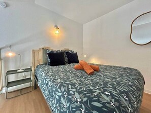 Een slaapkamer met een bed, kussens, een nachtkastje en een spiegel aan de muur.