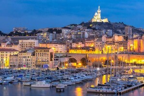Un port de plaisance où sont amarrés de nombreux bateaux, un bâtiment imposant surmonté d’une tour, situé sur une colline, et des bâtiments urbains environnants.