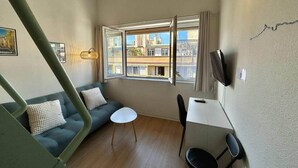 Una habitación pequeña con un sofá, una mesa y una silla. Hay una ventana que da a edificios.