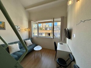 Una habitació petita amb un sofà, una taula i una cadira. Hi ha una finestra amb vista a edificis.