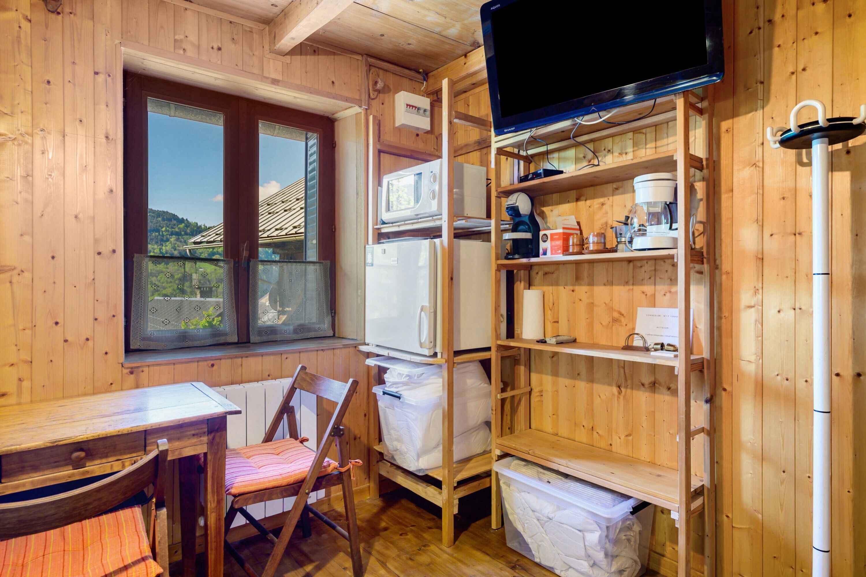 L’intérieur d’un chalet en bois comprend une table à manger, une chaise, une fenêtre offrant une vue, des étagères intégrées avec des appareils de cuisine et un téléviseur.