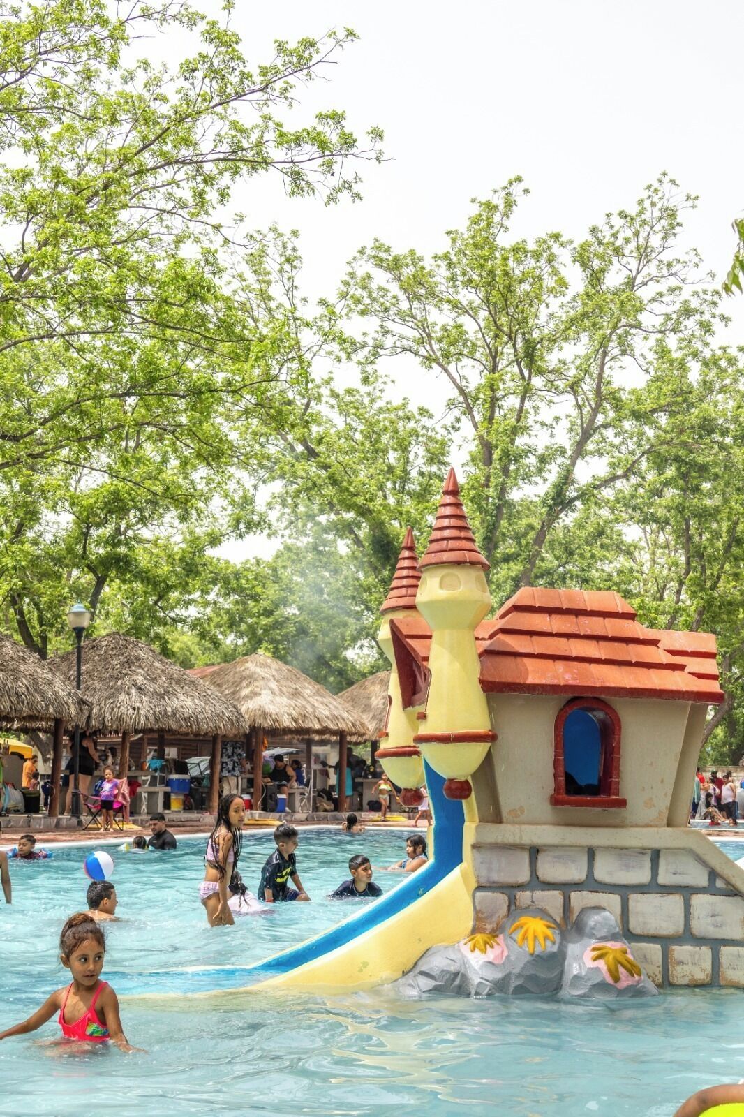 A swimming pool with a castle-like structure, surrounded by trees and thatched-roof huts.