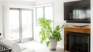 A modern living room with a white sofa, a large TV, a wooden ceiling beam, and a sliding glass door leading to a balcony.