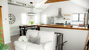 A modern kitchen with a central island, a dining area with a white sofa, and a visible clock on the wall.