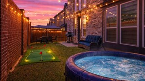 A backyard with a hot tub, string lights, a patio with chairs, and a small putting green. - Worldcup Ready Luxury 6BR  Game Room  EV  Pets OK  Sleeps 24 (Irving)
