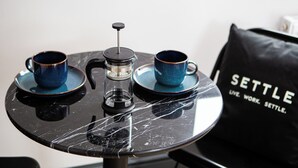 Una mesa con dos tazas azules y una cafetera, una silla negra y un bolso negro con el texto 'SETTLE'.