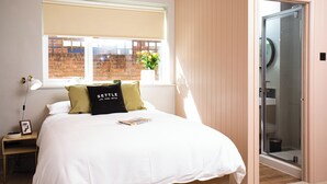 Un dormitorio con una cama, una mesita de noche, una lámpara, una ventana que da a un edificio de ladrillo y una puerta al baño.
