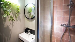 Un baño con lavamanos blanco, un espejo, ducha con cabezal de ducha de mano y plantas verdes en la pared.