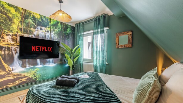 A bedroom with a large wall mural, a flat-screen TV, a bed with a green blanket, and a potted plant. - Large apartment for 8 people, Sauna & SPA bath, travel around the world (Drusenheim)