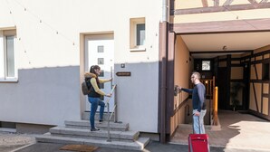 A building entrance with steps leading up to a door, a sign above the door, and a person with a red suitcase. - Large apartment for 8 people, Sauna & SPA bath, travel around the world (Drusenheim)