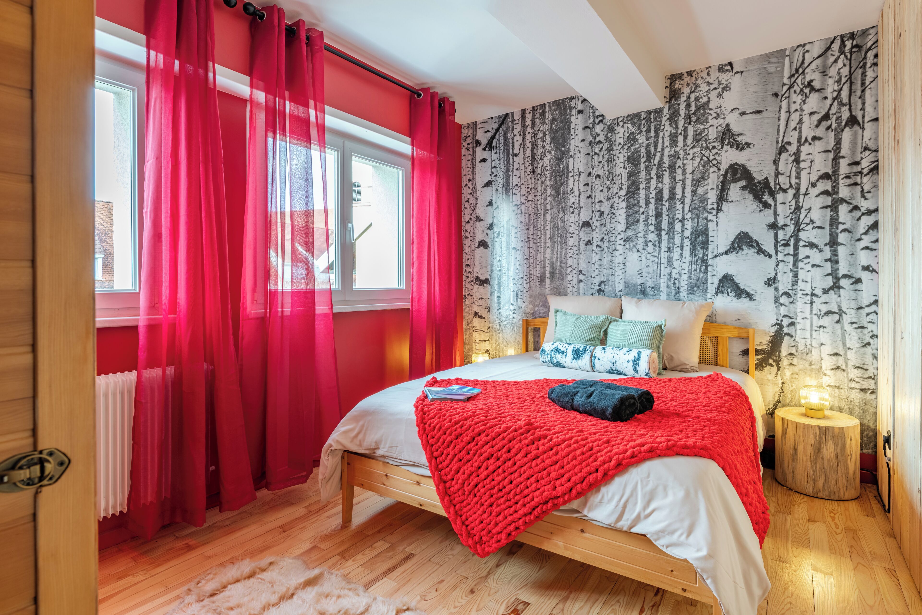 A bedroom with a wooden bed, red curtains, a birch tree wallpaper, and a small bedside lamp.