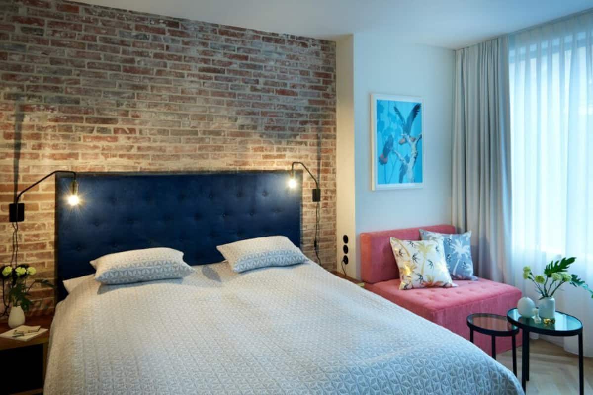 A hotel room with a brick wall, a large bed with a dark headboard, a pink sofa, and a small table with a vase of flowers.