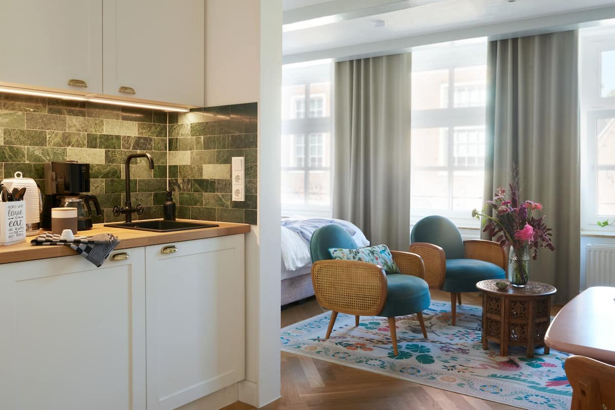 A modern kitchen with a tiled backsplash, a coffee machine, and a sink. There is a bed with a blue headboard and two wicker chairs in a living area with a view of buildings through large windows.