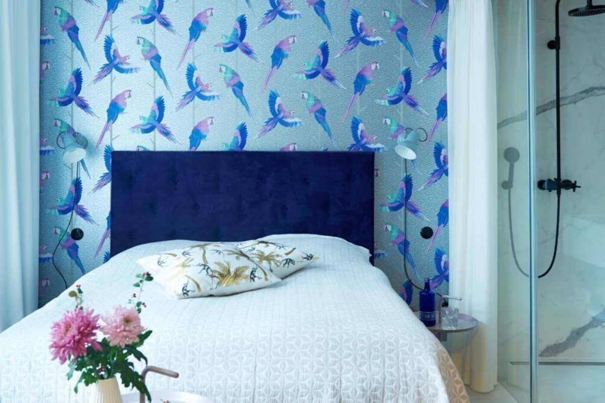 A bedroom with a patterned wallpaper, a dark headboard, a white bedspread, and a nightstand with a vase of flowers.