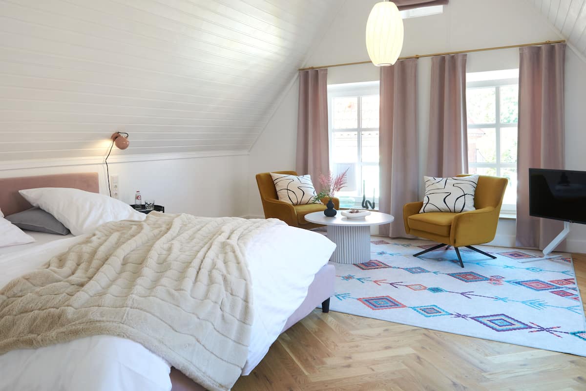 A hotel room with a bed, two armchairs, a small table, a TV, and a window with curtains.