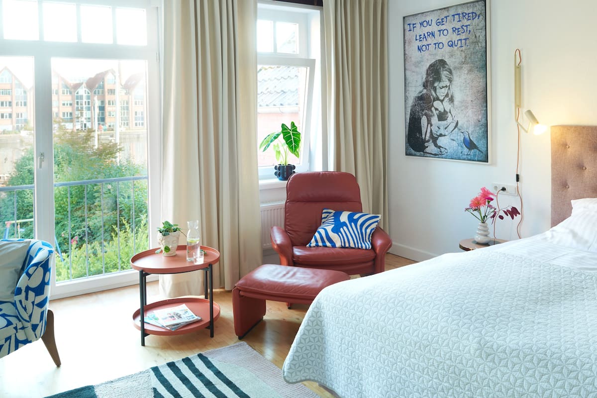 A hotel room with a bed, a red leather chair, a small table with a vase of flowers, a window with curtains, and a view of buildings and greenery.