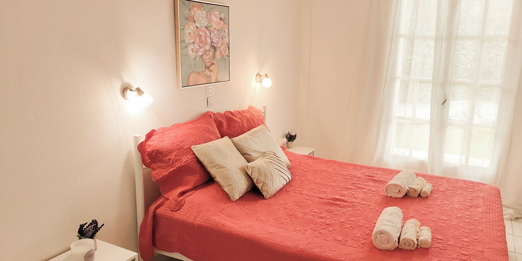 A neatly made bed with a red bedspread, pillows, and rolled towels on a white headboard.