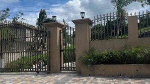 Uma entrada fechada por grades, com pavimento de tijolos estampados, portões de metal e um muro com detalhes decorativos.