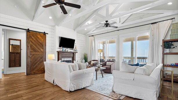 Un salon spacieux, meublé de blanc, avec un plancher en bois, un ventilateur de plafond et une grande fenêtre donnant sur un beau paysage.