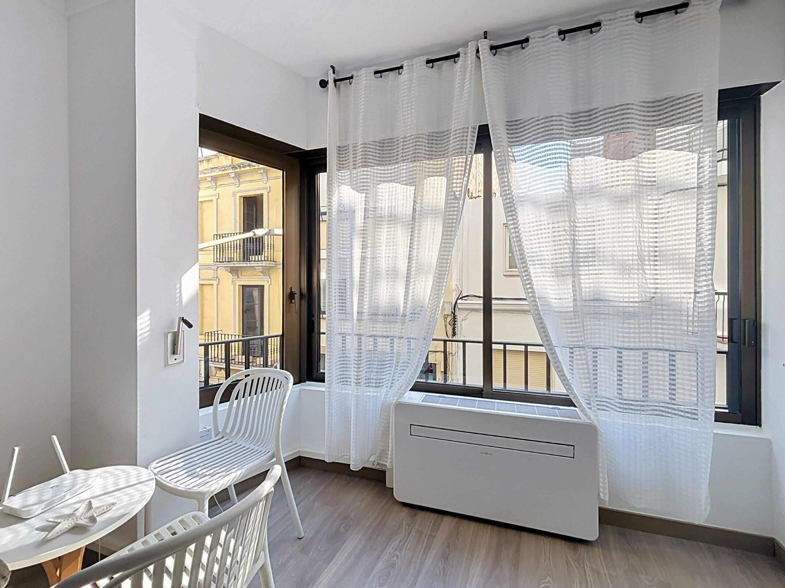Ein Balkon mit weißem Stuhl, einem kleinen Tisch und einem Fenster mit weißen Vorhängen.