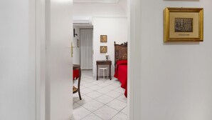 Ein Flur mit weiß gefliestem Boden, einer Holztür, einem kleinen Tisch mit Stuhl und einem roten Sofa.