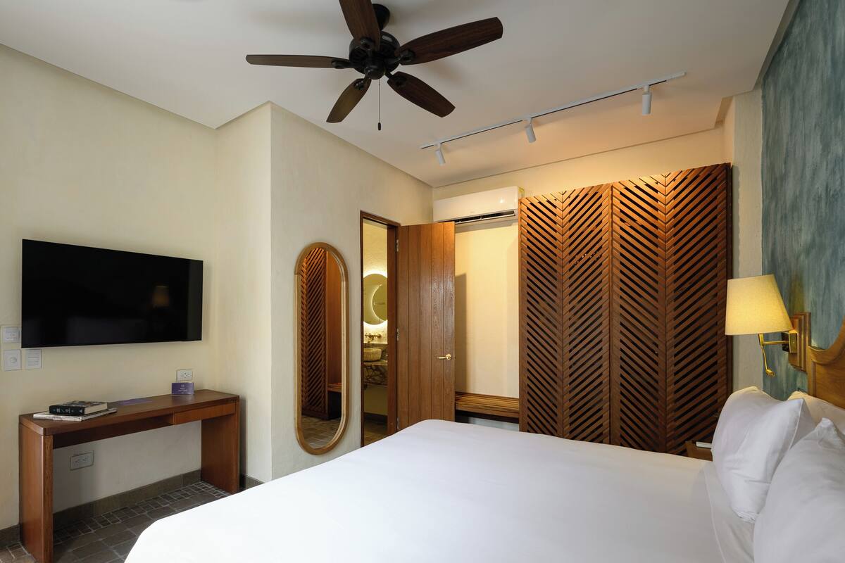 Habitación de hotel con cama, escritorio, televisor, ventilador de techo, espejo, armario y lámpara.