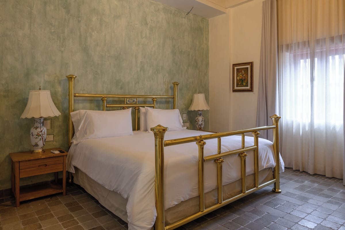 Una habitación con una cama, dos mesitas de noche, una lámpara de noche, un cuadro y una ventana con cortinas.
