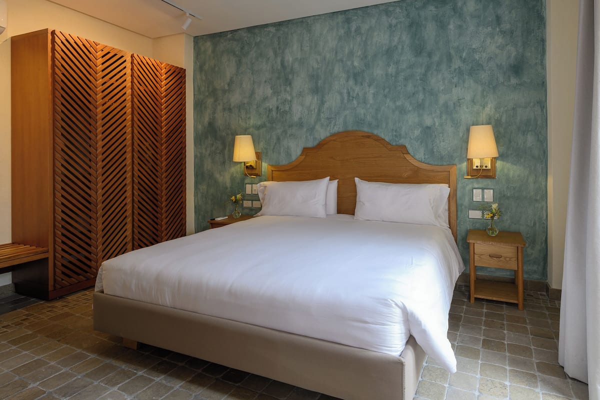 Una habitación de hotel con una cama grande, cabecera de madera, dos mesitas de noche con lámparas, una luz empotrada en la pared y un panel de madera con diseño.