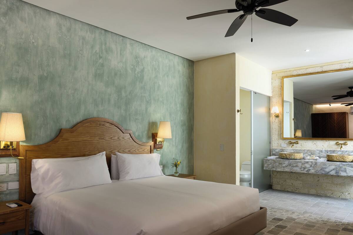 Habitación de hotel con cama, lámparas de noche, ventilador de techo y baño con lavamanos y espejo.