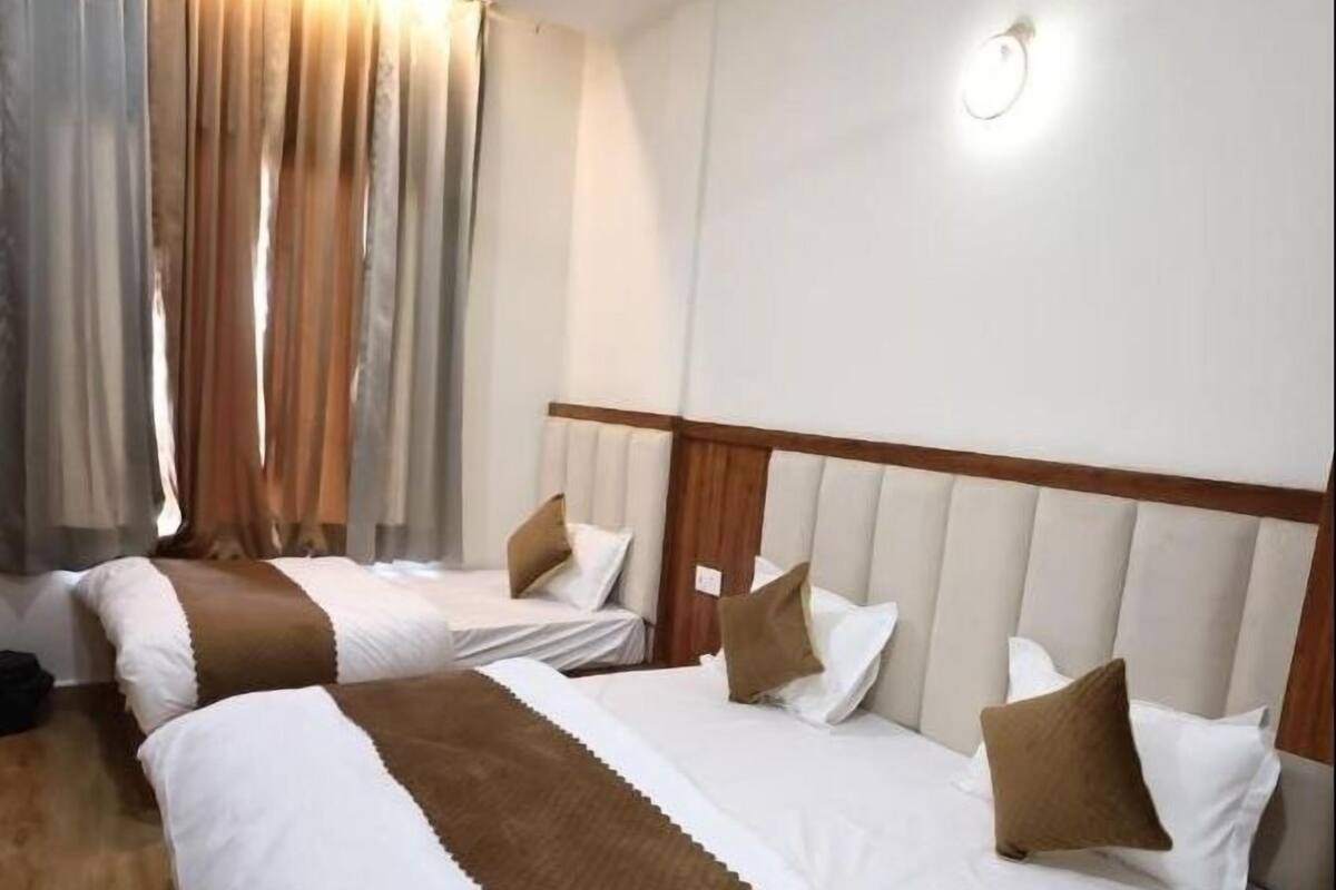 A hotel room with two beds, a wooden floor, a large window with curtains, and a wall-mounted light.