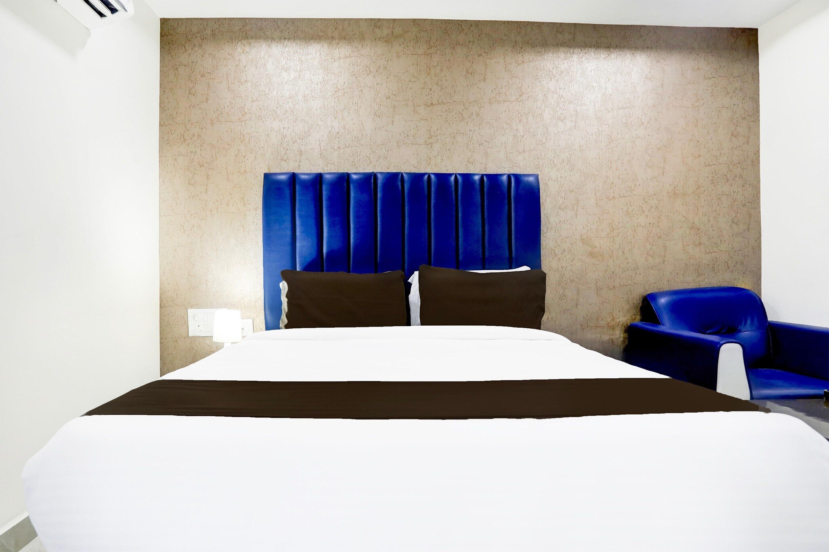 A hotel room with a large bed, a blue headboard, a blue armchair, and a wall-mounted air conditioning unit.