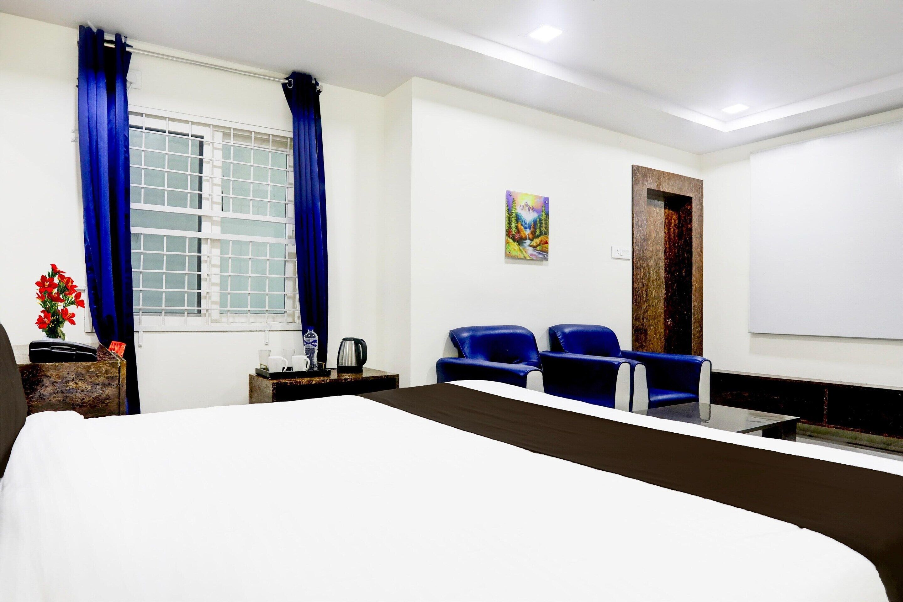 Habitación de hotel con cama, dos sillas azules, un cuadro en la pared y una ventana con cortinas azules.