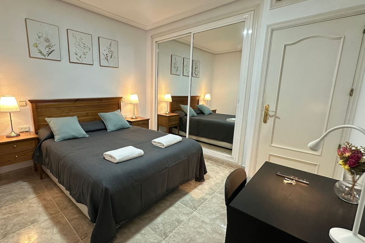 A hotel room with a bed, bedside tables, a desk, a mirror, and framed floral artwork on the wall.