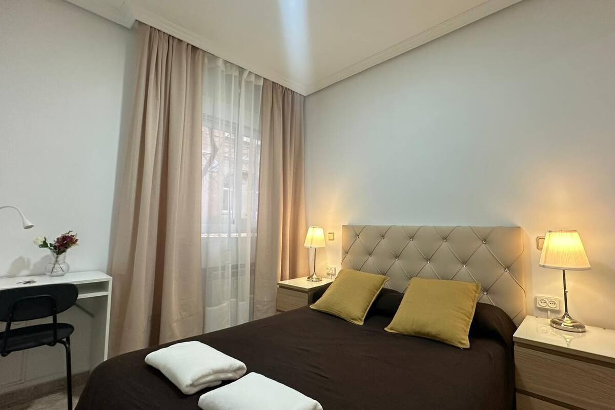 A hotel room with a bed, two bedside lamps, a desk with a chair, a window with curtains, and a vase with flowers.