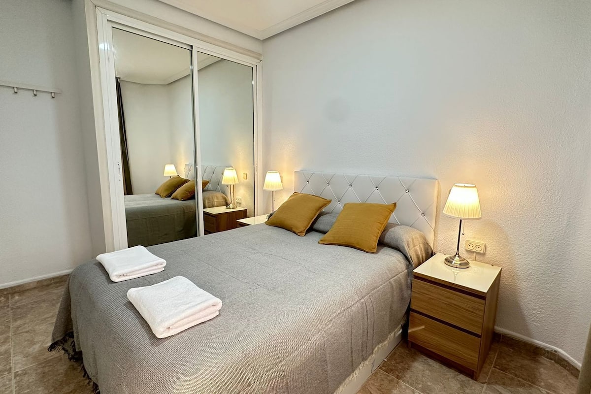 A neatly made bed with pillows, a bedside table with a lamp, a mirror, and a wardrobe.