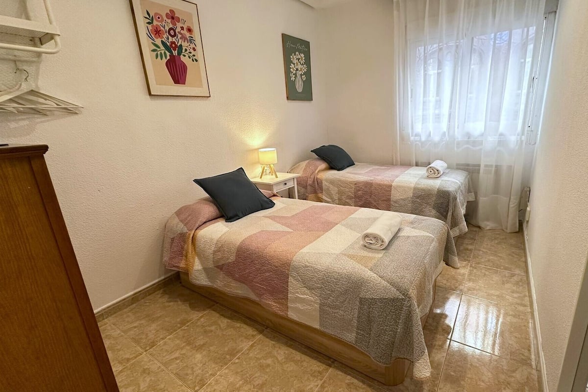 A hotel room with two beds, a wooden dresser, a lamp, and a window with sheer curtains.