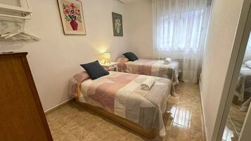 Una habitación de hotel con dos camas, un aparador de madera, una lámpara y una ventana con cortinas translúcidas.