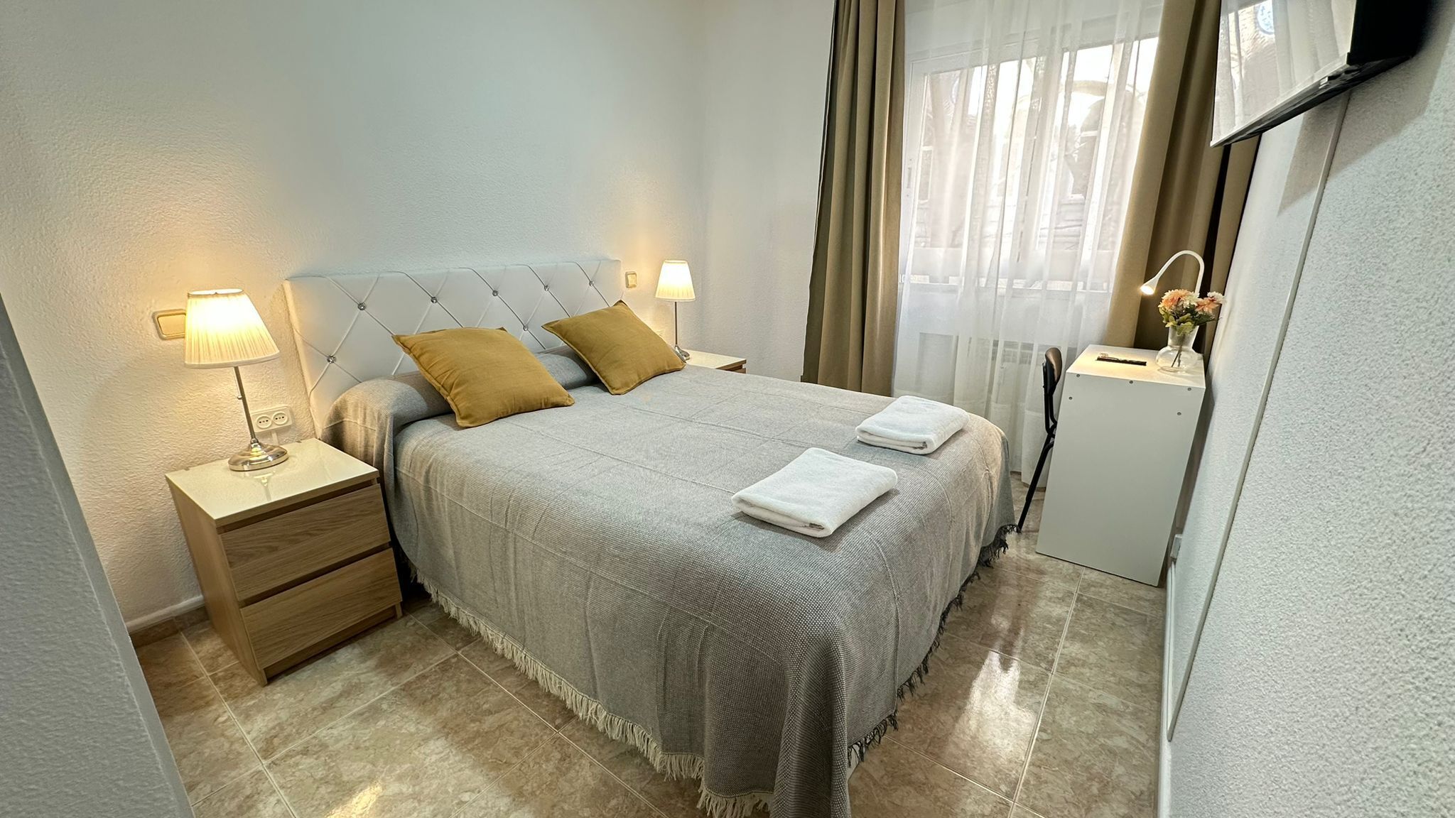 A hotel room with a bed, two bedside lamps, a nightstand, a dresser, a small table with a lamp, a mirror, a radiator, and a window with curtains.