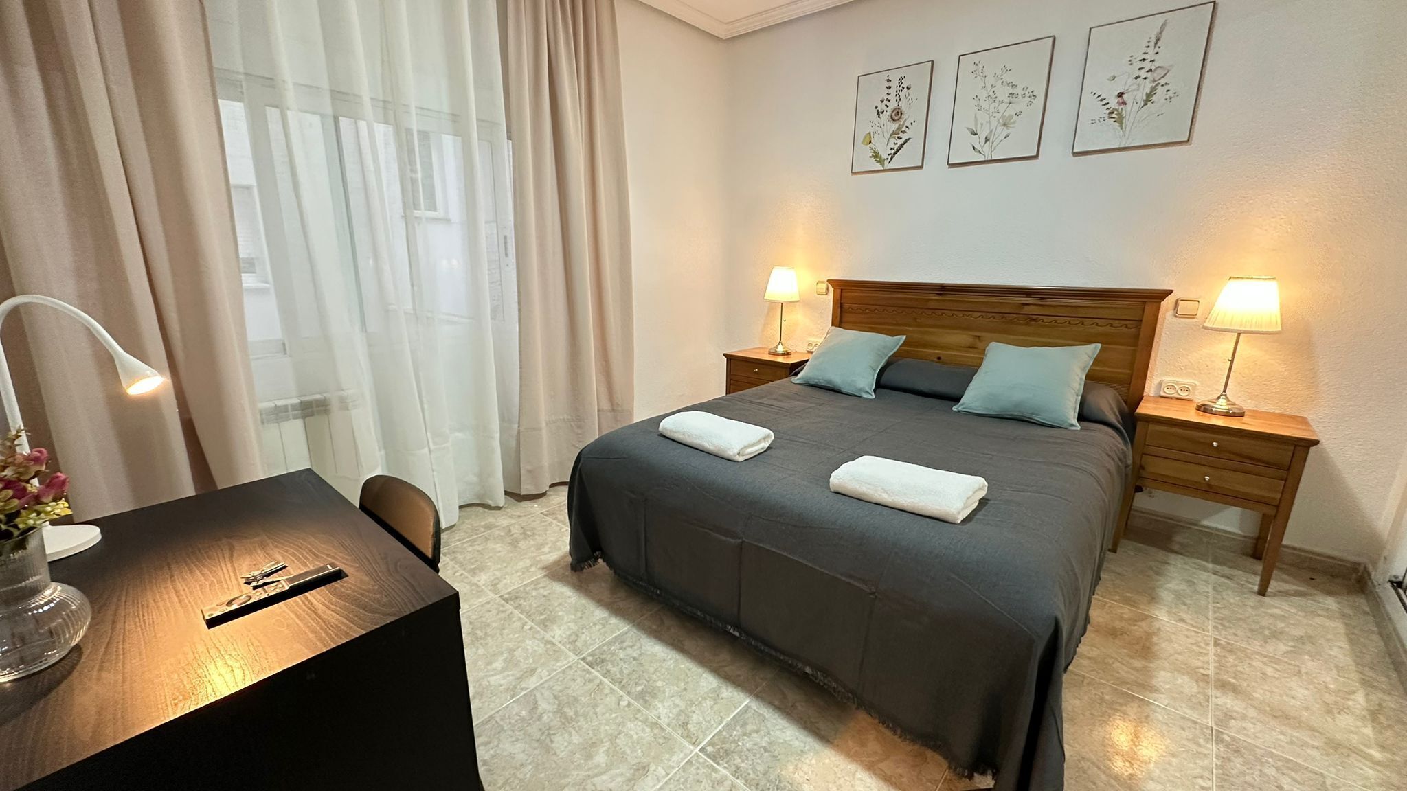 A hotel room with a bed, two bedside tables with lamps, a desk with a chair, and a window with curtains.