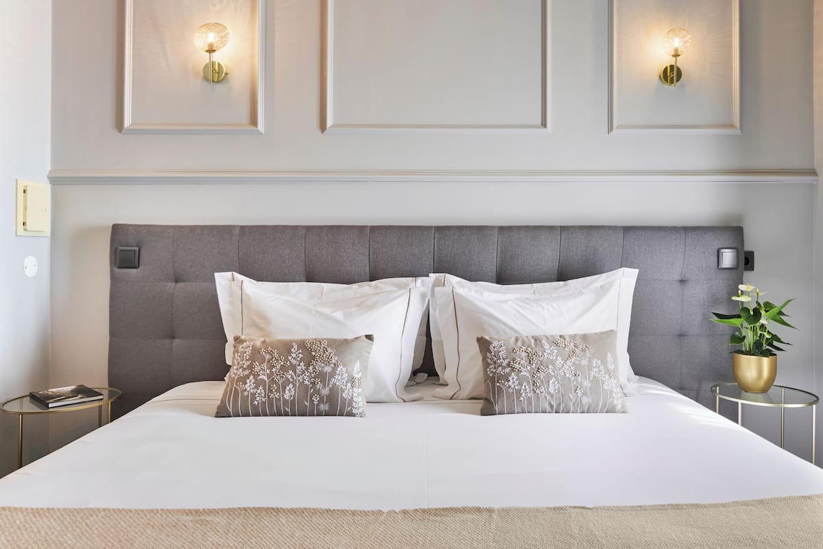 A neatly made bed with a grey upholstered headboard, two bedside tables with lamps, and a potted plant on the right.