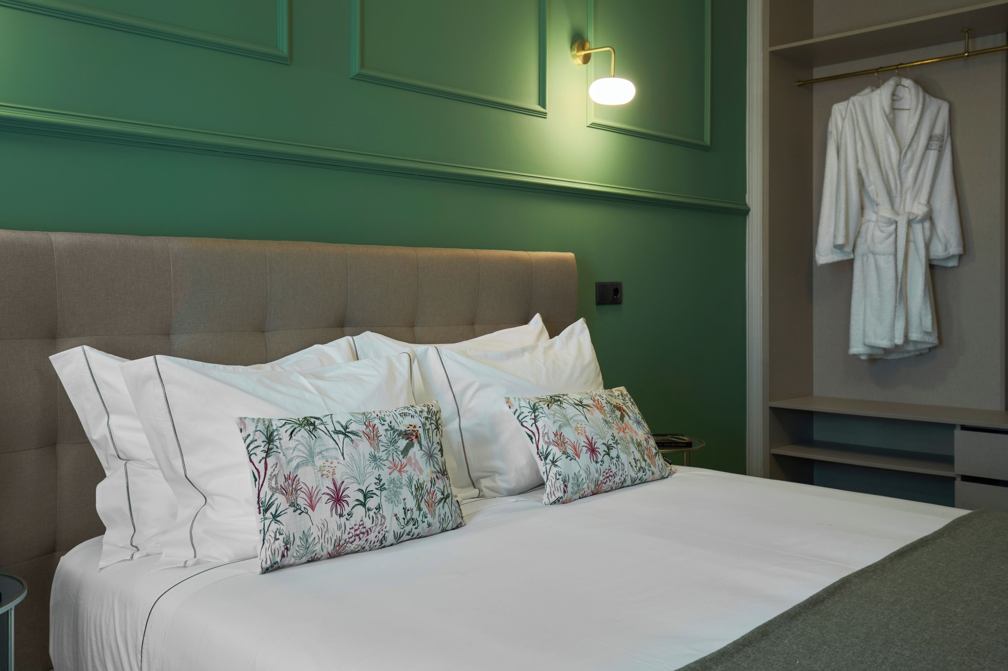 A neatly made bed with white bedding and floral pillows, a green wall with a light fixture, and a robe hanging on a rack.